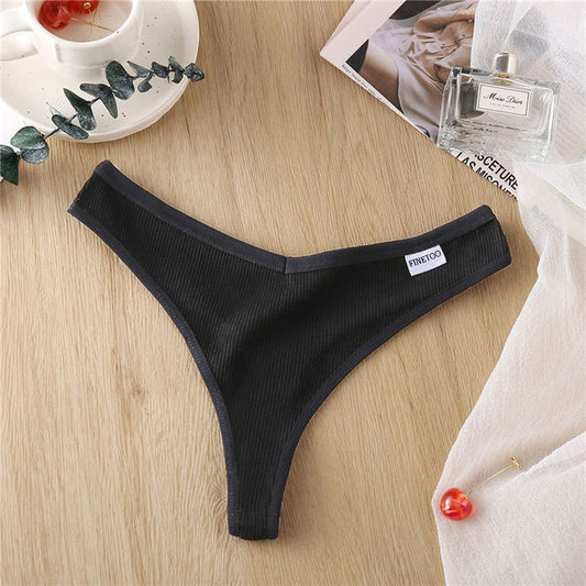 Black thong on a wooden surface with a cup, plant, and magazine in the background.